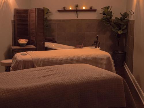 A serene spa room with two massage tables, soft lighting, and a bathtub in the background, surrounded by plants.