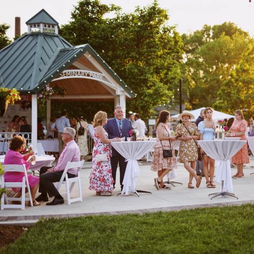 Guests socialize at an outdoor event in a park, enjoying drinks and conversation near a gazebo.