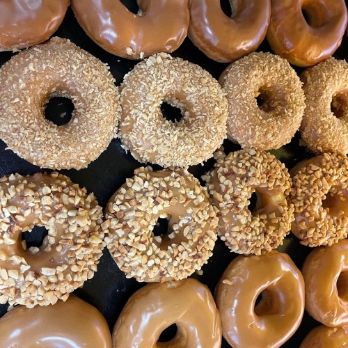 A variety of donuts, including glazed, nut-covered, and crumb-topped, arranged in rows on a black surface.