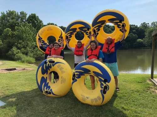 A group of five people in life jackets holding large yellow inner tubes by a lake, surrounded by greenery.