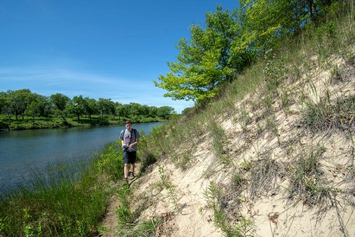 A person walks along a sandy riverbank with lush greenery and blue sky in the background.