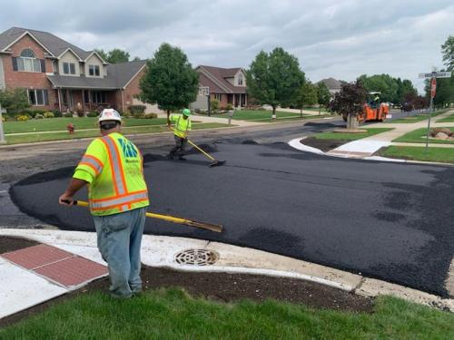 Two workers in reflective vests smooth freshly laid asphalt on a residential street corner.