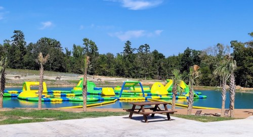 A colorful inflatable water park on a lake, surrounded by trees and a picnic table in the foreground.