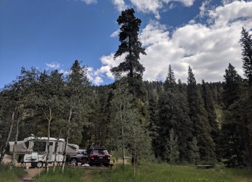 A campsite surrounded by tall trees, featuring a pop-up camper and a parked car under a blue sky with clouds.