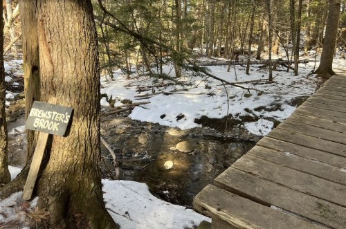 A wooden sign reading "Brewster's Brook" next to a snowy stream and a wooden bridge in a forested area.