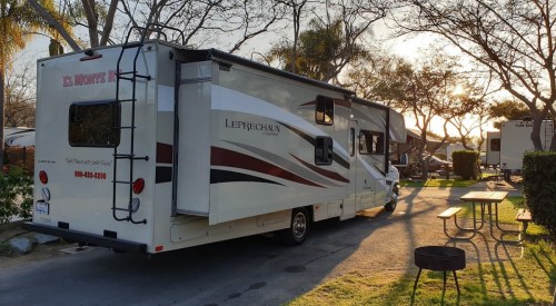 A parked RV on a tree-lined road, with picnic tables nearby and a sunset in the background.