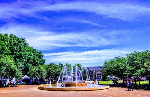 A fountain in a park surrounded by trees and people, under a bright blue sky with wispy clouds.