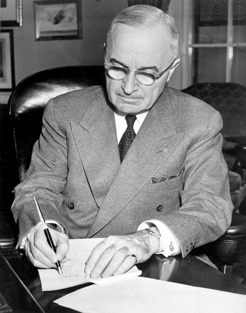A man in a suit and glasses writes on paper at a desk, focused on his work.