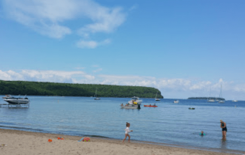 A sunny beach scene with boats on the water, a child playing in the sand, and lush green hills in the background.