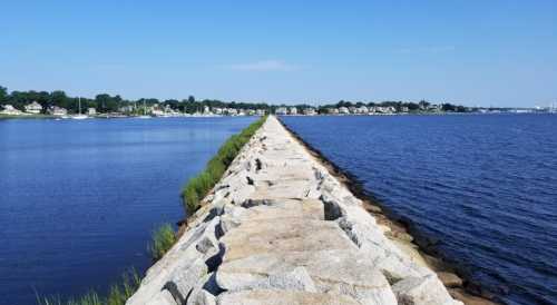 A stone jetty extends into a calm body of water, with a clear blue sky and houses visible in the background.