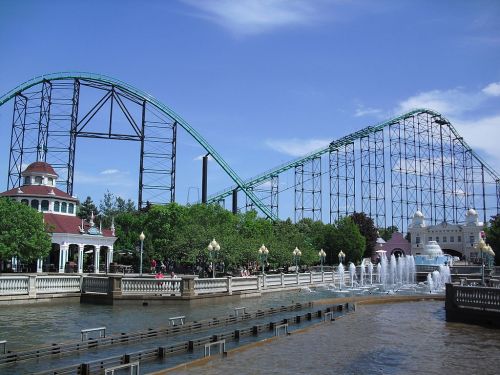 A roller coaster towers over a park with a fountain and greenery, under a clear blue sky.