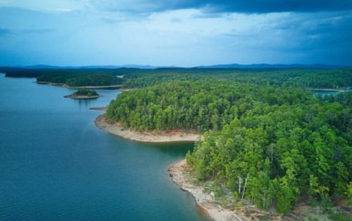 Aerial view of a serene lake surrounded by lush green forests and distant hills under a cloudy sky.