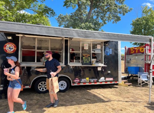 A food truck with people ordering and enjoying food at an outdoor event on a sunny day.