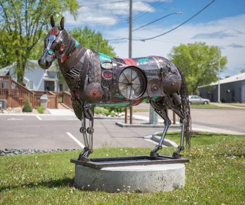 A metal sculpture of a horse made from various recycled materials, standing on a circular base in a grassy area.