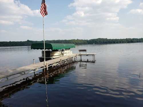 A peaceful lakeside scene featuring a dock, a boat with a green canopy, and an American flag waving in the breeze.