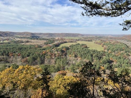 A scenic view of rolling hills and forests in autumn, with colorful foliage under a partly cloudy sky.