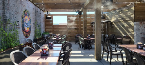 Outdoor dining area with wooden tables, chairs, and misting fans, surrounded by greenery and wooden walls.