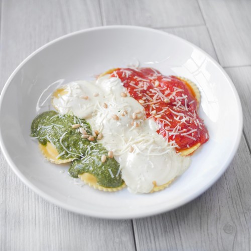A bowl of ravioli topped with green pesto, red sauce, and creamy white sauce, garnished with pine nuts and cheese.