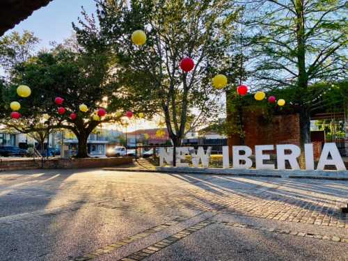 A sunny plaza in New Iberia, decorated with red and yellow lanterns hanging from trees. Large "NEW IBERIA" letters in foreground.