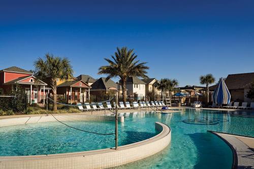 A sunny pool area with palm trees, lounge chairs, and colorful houses in the background under a clear blue sky.