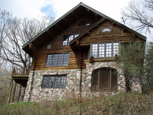 A rustic log cabin with stone accents, large windows, and a balcony, surrounded by trees and a cloudy sky.