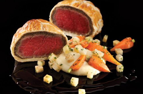 A plated dish featuring beef Wellington, sliced to reveal pink meat, served with mashed potatoes and colorful vegetables.