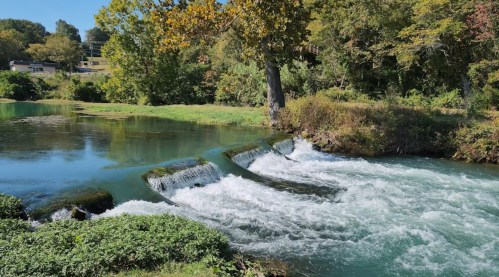 A serene river scene with gentle waterfalls, surrounded by lush greenery and trees under a clear blue sky.