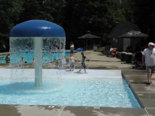 Children play near a water feature at a pool, with adults and a shaded area in the background.