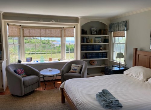 Cozy bedroom with a bed, two armchairs, a small table, and a view of greenery and water through large windows.
