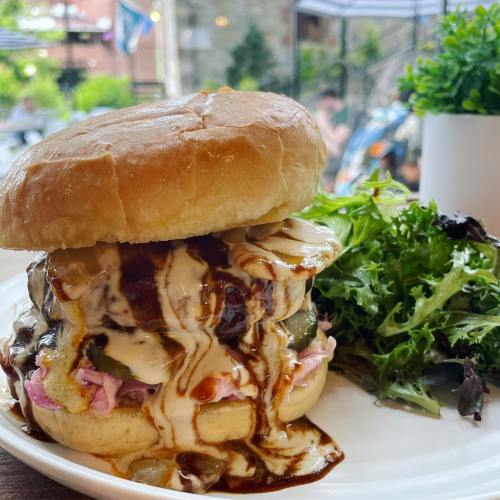 A close-up of a juicy burger with sauce, pickles, and slaw, served with a side of fresh greens on a white plate.