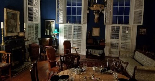 A dimly lit room with blue walls, antique furniture, and a dining table set for a meal, featuring a chandelier and artwork.