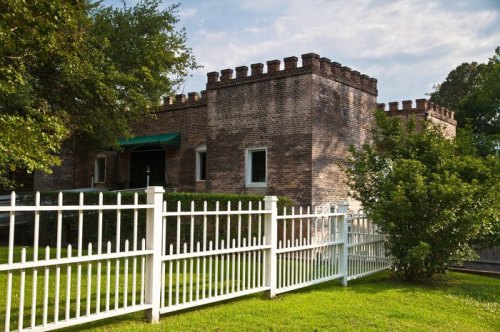A brick building with a castle-like design, surrounded by a white picket fence and green grass.