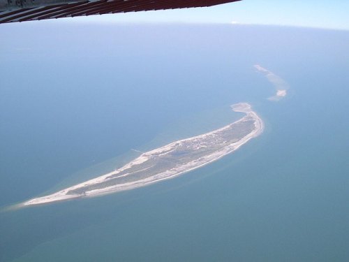 Aerial view of a long, narrow island surrounded by blue water, with sandy beaches and greenery visible.