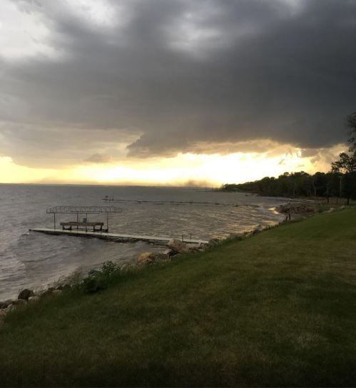 A stormy sky looms over a calm lake, with a grassy shore and a wooden dock extending into the water.