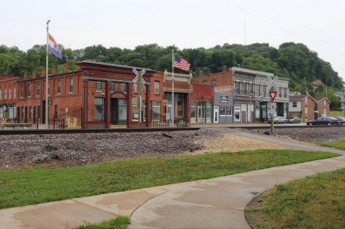 Historic buildings line a street near a railroad track, with flags and greenery in the background.