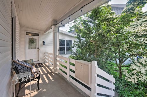 A cozy porch with a bench, mailbox, and greenery, leading to a white house with large windows.