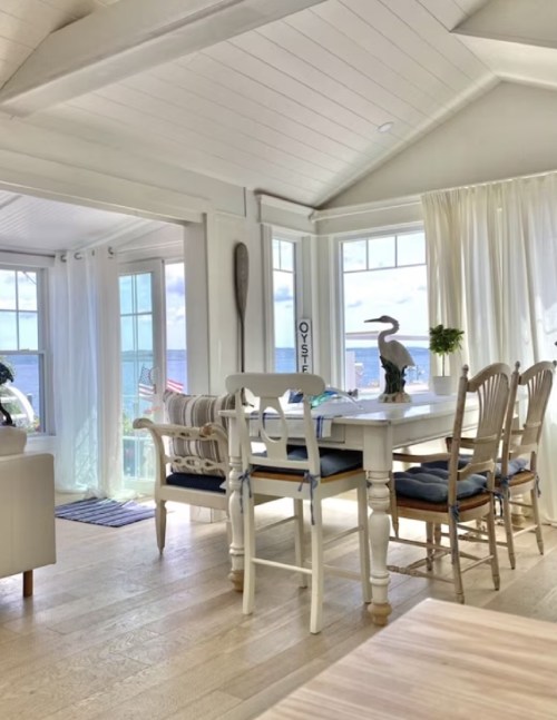 Bright coastal dining area with a white table, chairs, and large windows showcasing a view of the ocean.