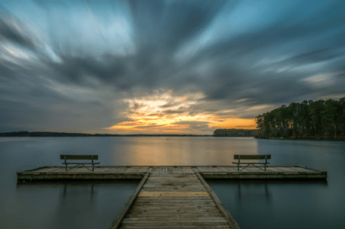 A serene lake view at sunset, featuring a wooden dock with two benches and dramatic clouds in the sky.