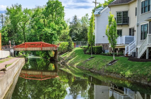 A serene canal scene with a red bridge, lush greenery, and modern homes along the water's edge.