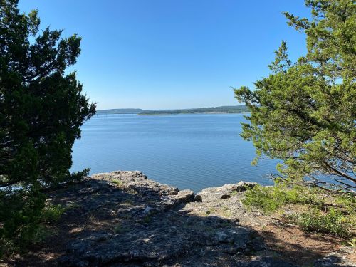 A serene view of a calm lake surrounded by trees and rocky terrain under a clear blue sky.