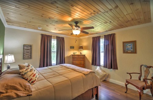 Cozy bedroom with wooden ceiling, a bed, dresser, and windows with curtains, featuring warm lighting and a chair.