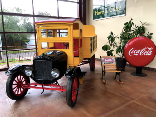 A vintage yellow delivery truck next to a red Coca-Cola sign and a small chair, set in a bright indoor space.