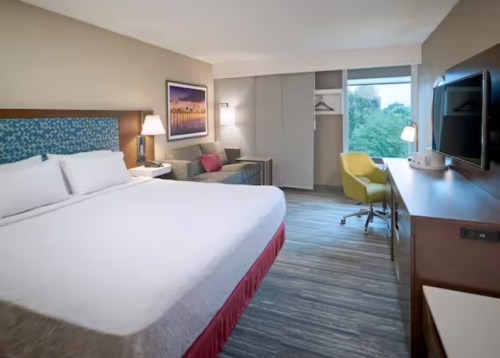 Modern hotel room featuring a king bed, seating area, desk, and large window with a view.