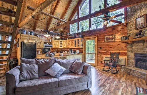 Cozy cabin interior with wooden beams, a sofa, kitchen, and large windows overlooking a forested area.