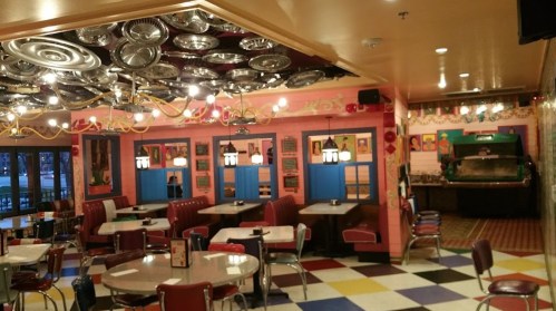 Colorful diner interior with retro decor, checkered floor, and booths, featuring vibrant artwork and hanging light fixtures.