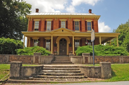 Historic brick house with a porch, surrounded by greenery and steps leading to the entrance, under a blue sky.