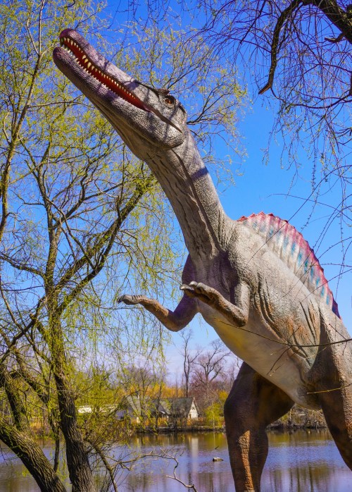 A large dinosaur statue with a long neck and colorful back stands near a lake, surrounded by trees and blue sky.