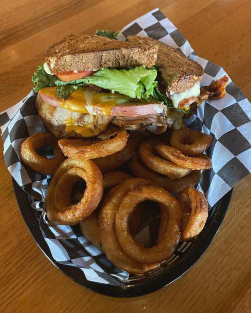 A hearty sandwich with lettuce, tomato, and cheese, served with a side of crispy onion rings in a black basket.