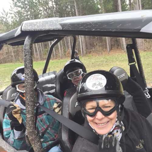 Three people wearing helmets and goggles smile while seated in a rugged off-road vehicle, surrounded by trees.