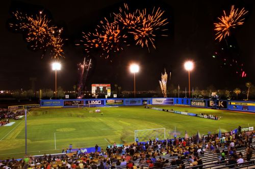 A lively soccer stadium at night, filled with fans, as fireworks light up the sky above the field.